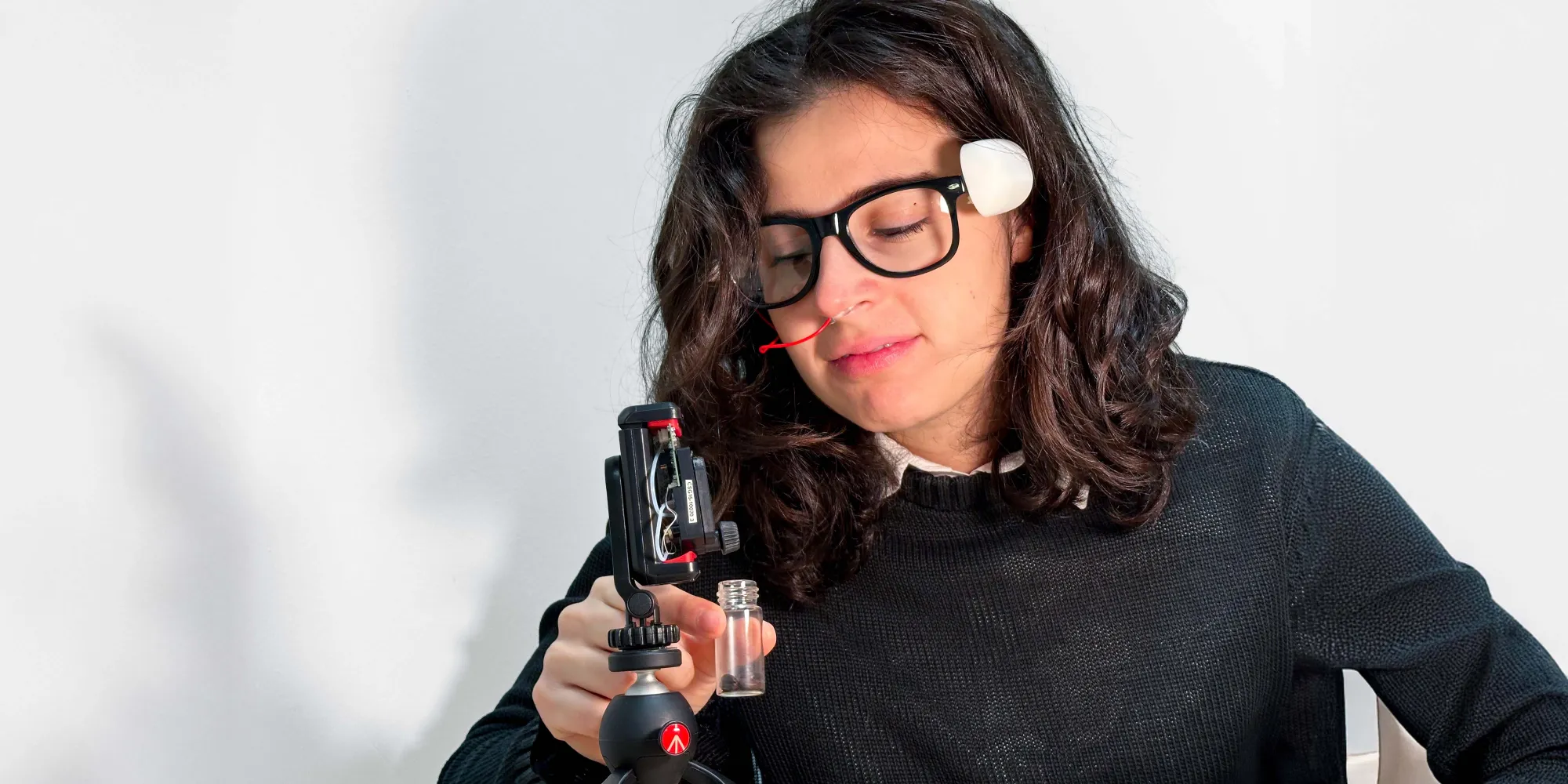 Person observing a small container under a technological device on a tripod, wearing a facial sensor.