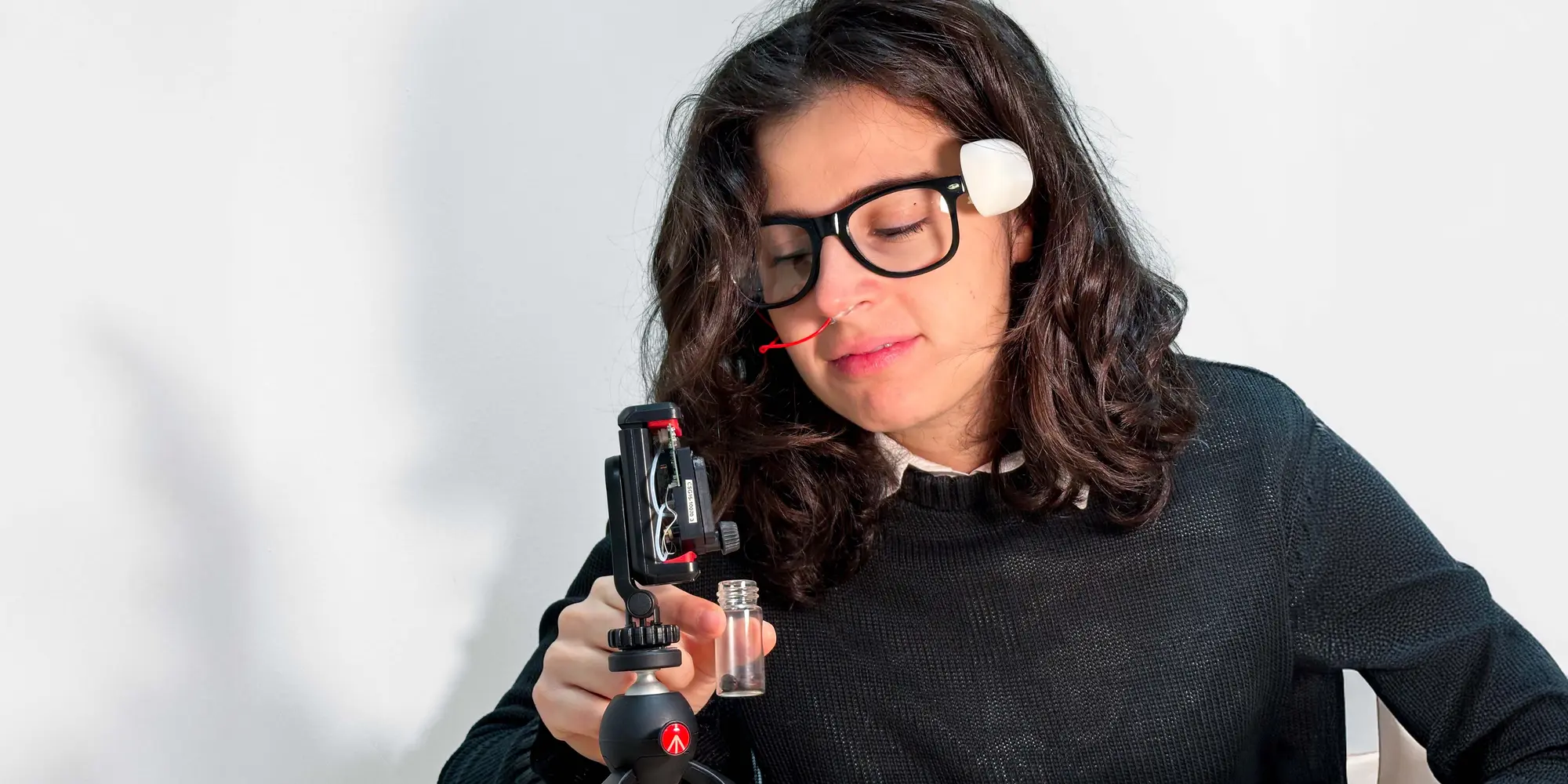Person observing a small container under a technological device on a tripod, wearing a facial sensor.