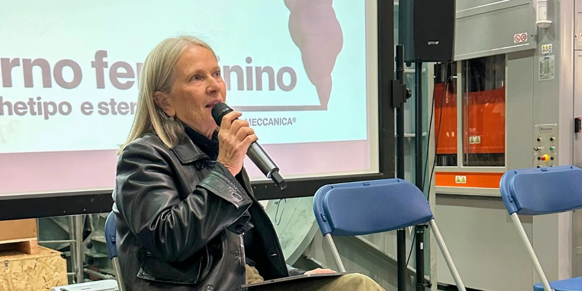Persona seduta parla al microfono davanti allo schermo dell’evento “Eterno Femminino” in un laboratorio industriale.