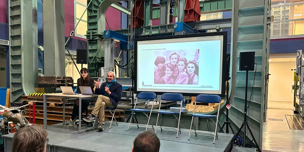 Relatore seduto parla davanti a una slide con un’immagine di gruppo femminile e il titolo sugli stereotipi, in un laboratorio industriale.