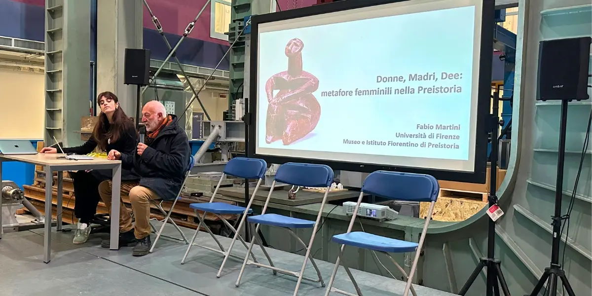 Relatore seduto parla davanti a una slide intitolata “Donne, Madri, Dee: metafore femminili nella Preistoria”, con una persona al computer accanto a lui in un ambiente di laboratorio industriale.