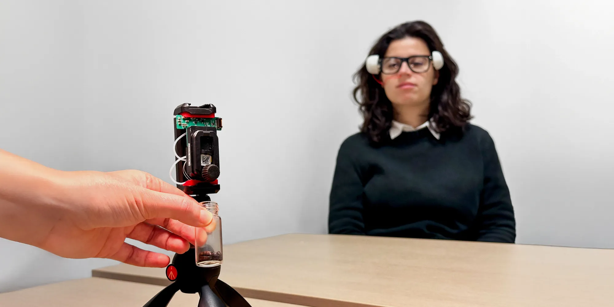 Hand placing a container under a technological device on a tripod, with a seated person in the background.