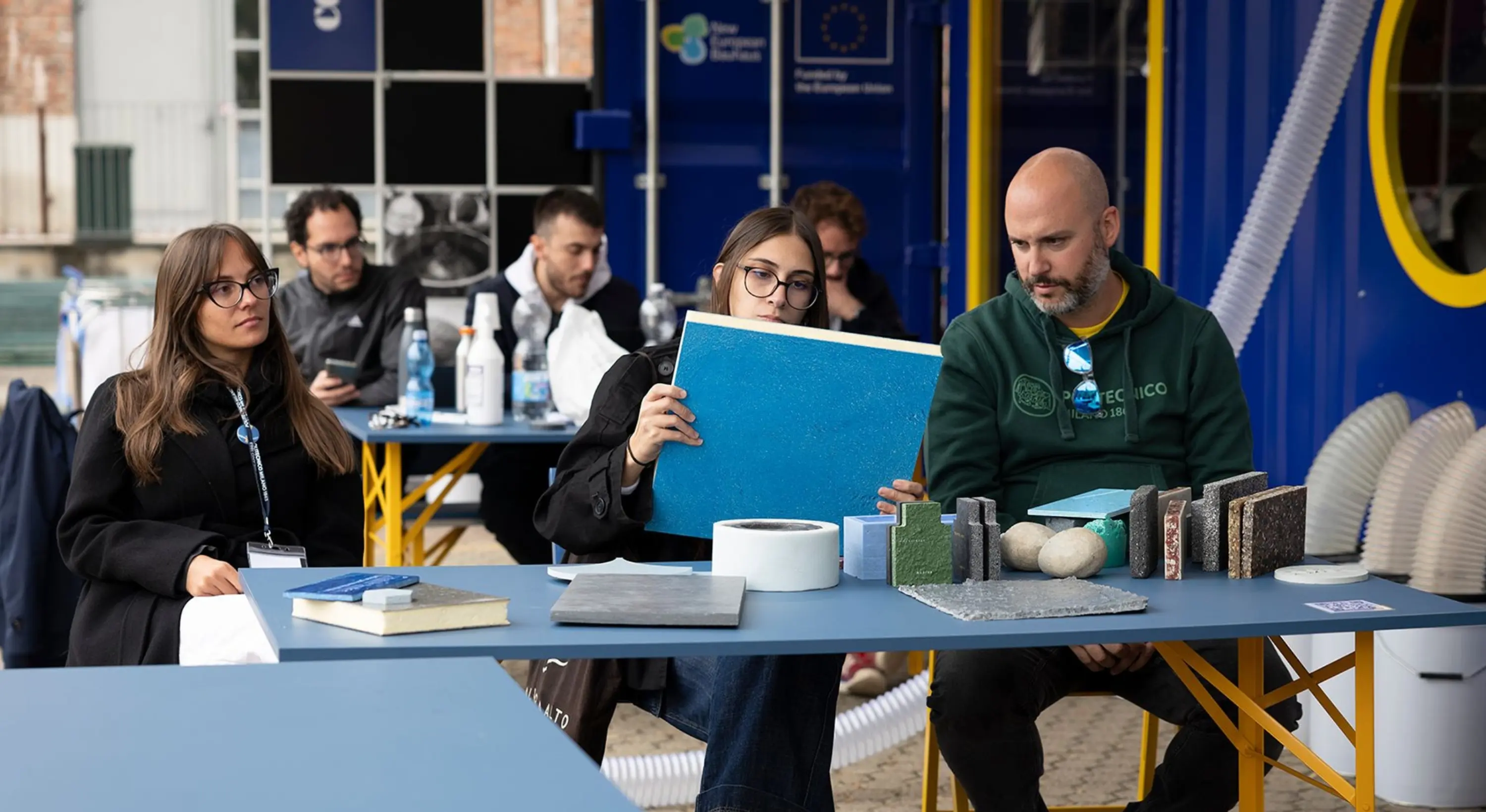 Persone sedute a un tavolo osservano e manipolano campioni di materiali, inclusi pannelli colorati e superfici testurizzate, durante un workshop all’aperto.