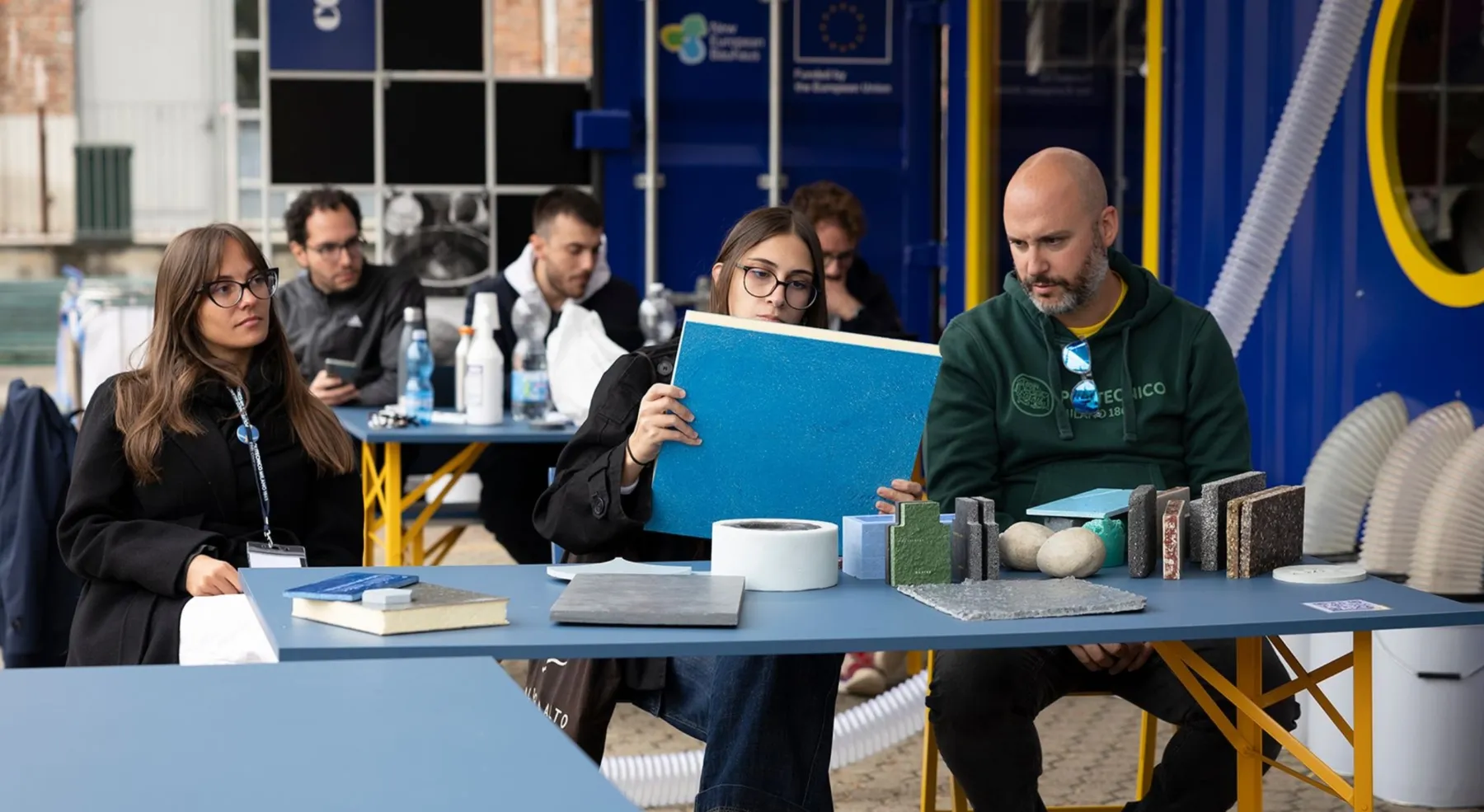 Persone sedute a un tavolo osservano e manipolano campioni di materiali, inclusi pannelli colorati e superfici testurizzate, durante un workshop all’aperto.