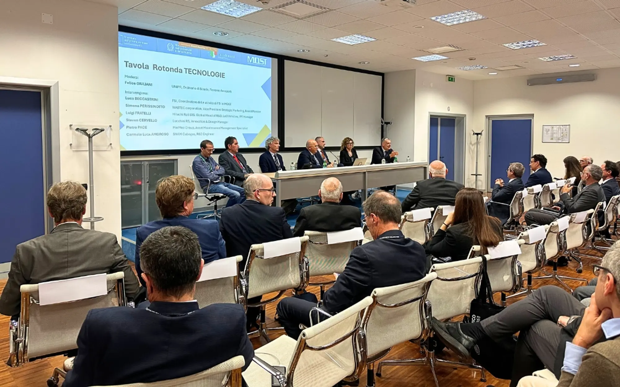 Audience attending a technology roundtable, with speakers seated in front of a screen showing the session title.