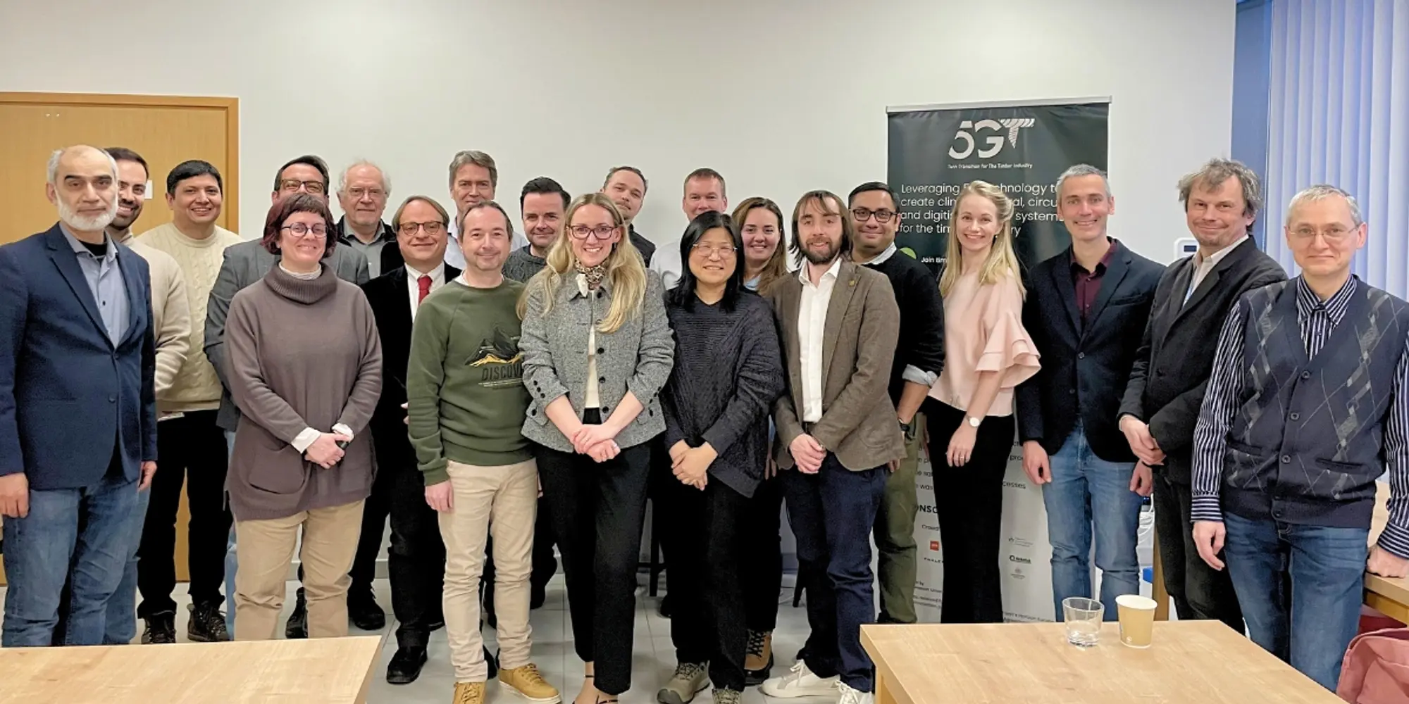 Group of people posing in front of a 5G-TIMBER project banner in a meeting room.