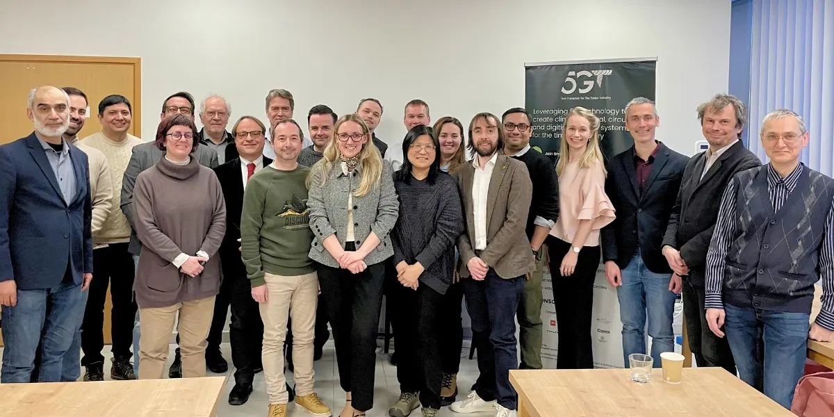 Group of people posing in front of a 5G-TIMBER project banner in a meeting room.
