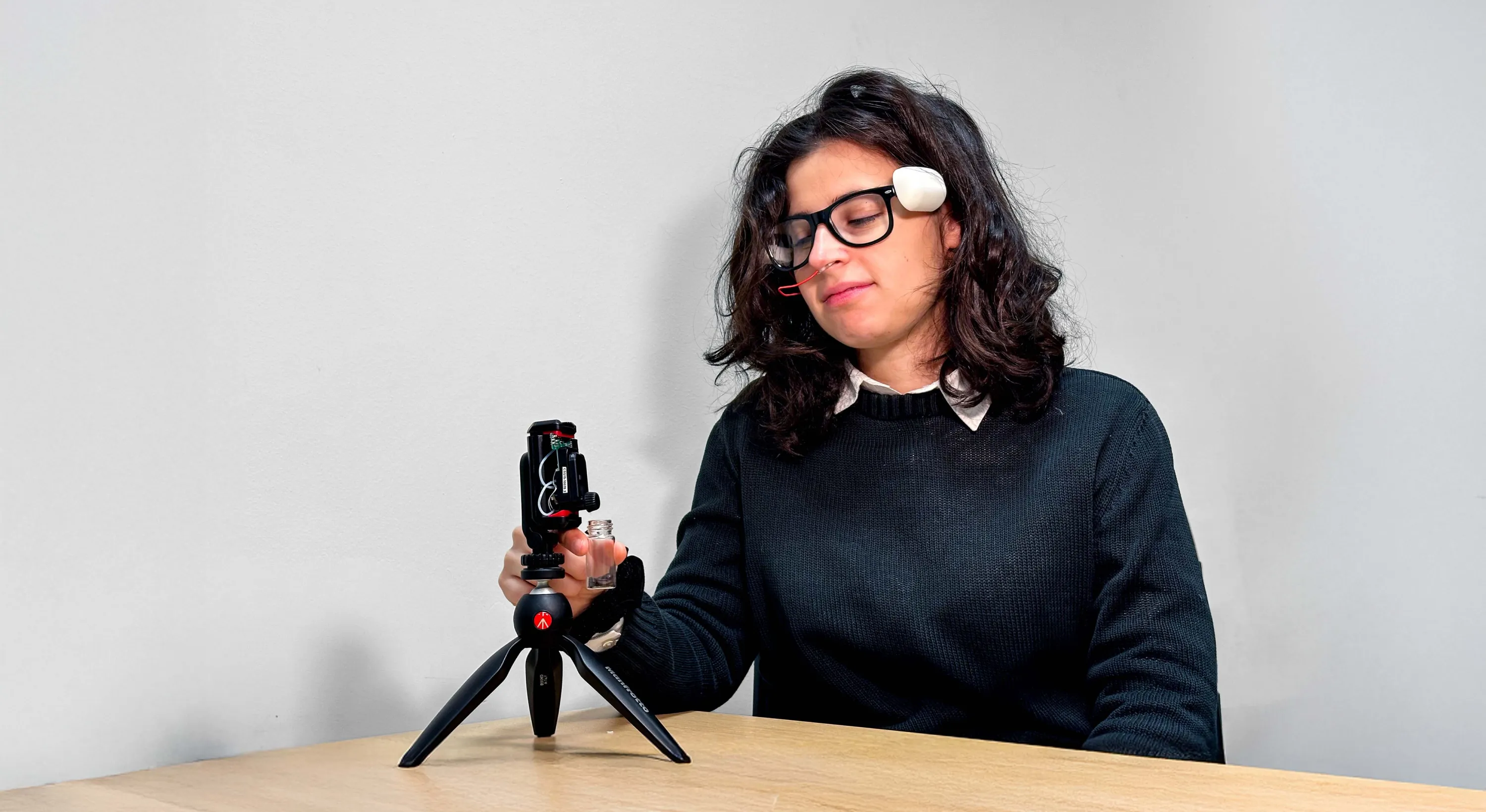 Person seated at a table using a technological device on a tripod and holding a small transparent container.