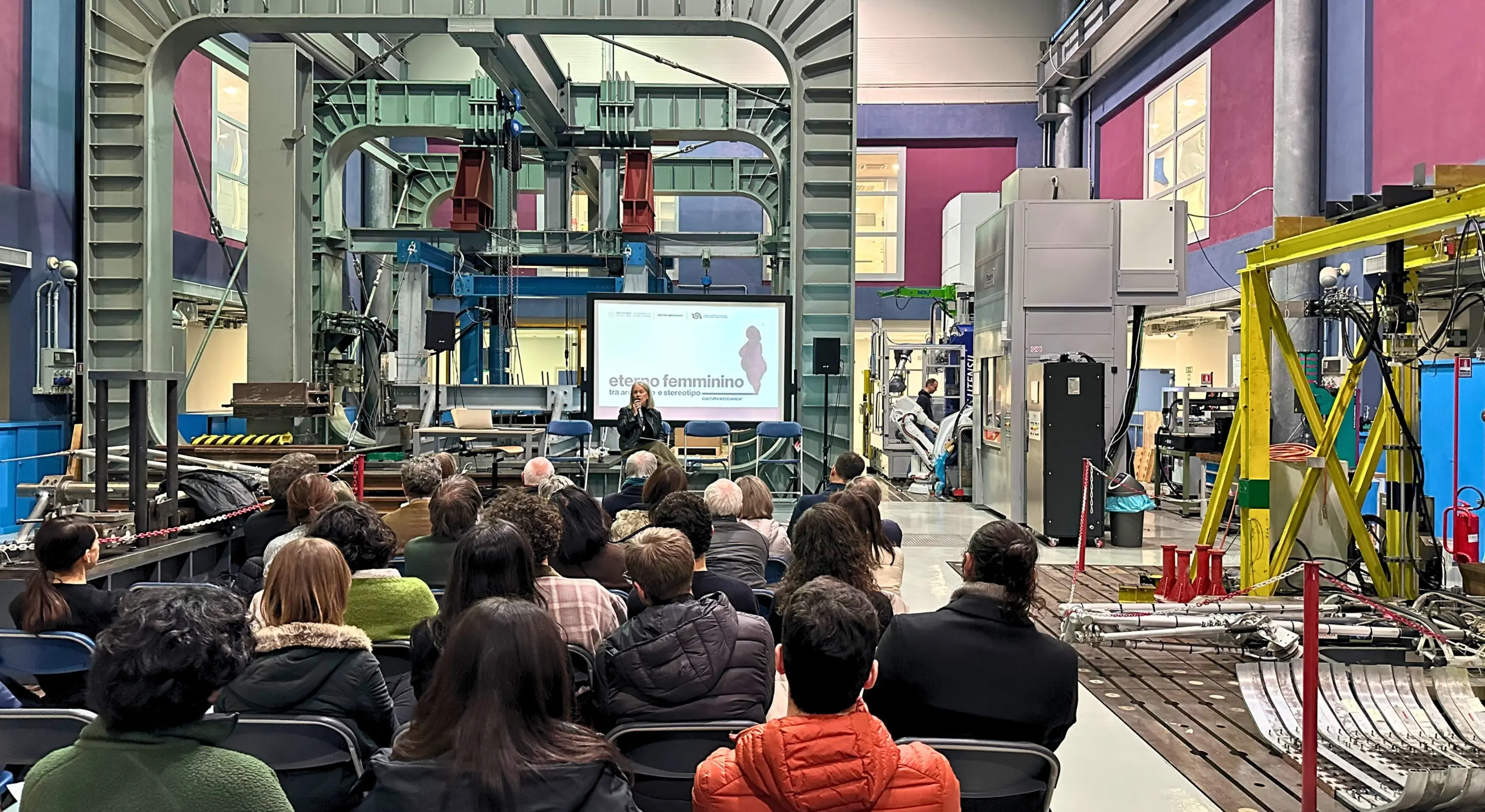 Pubblico seduto in un laboratorio industriale mentre una relatrice parla davanti a uno schermo con il titolo “Eterno Femminino”.