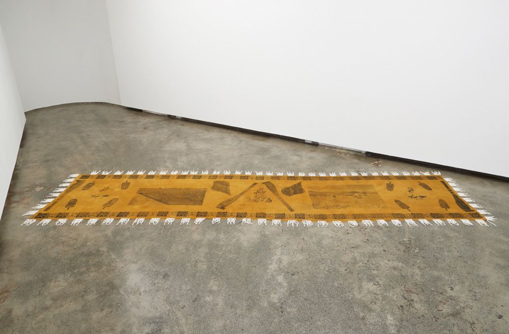 Image of hand-made textile by Quishile, made with assistance from her Aaji. 5 metre long cotton, dyed with genda (marigolds) and stitched with shells across all four sides of the textile work. Textile is screen printed and block printed with archival images of Girmit, Girmitiya women and motifs. The textile is lying on the ground and is surrounded by white walls. Textile work: Your Woman is a Very Bad Woman, shown at Firstdraft in 2018.