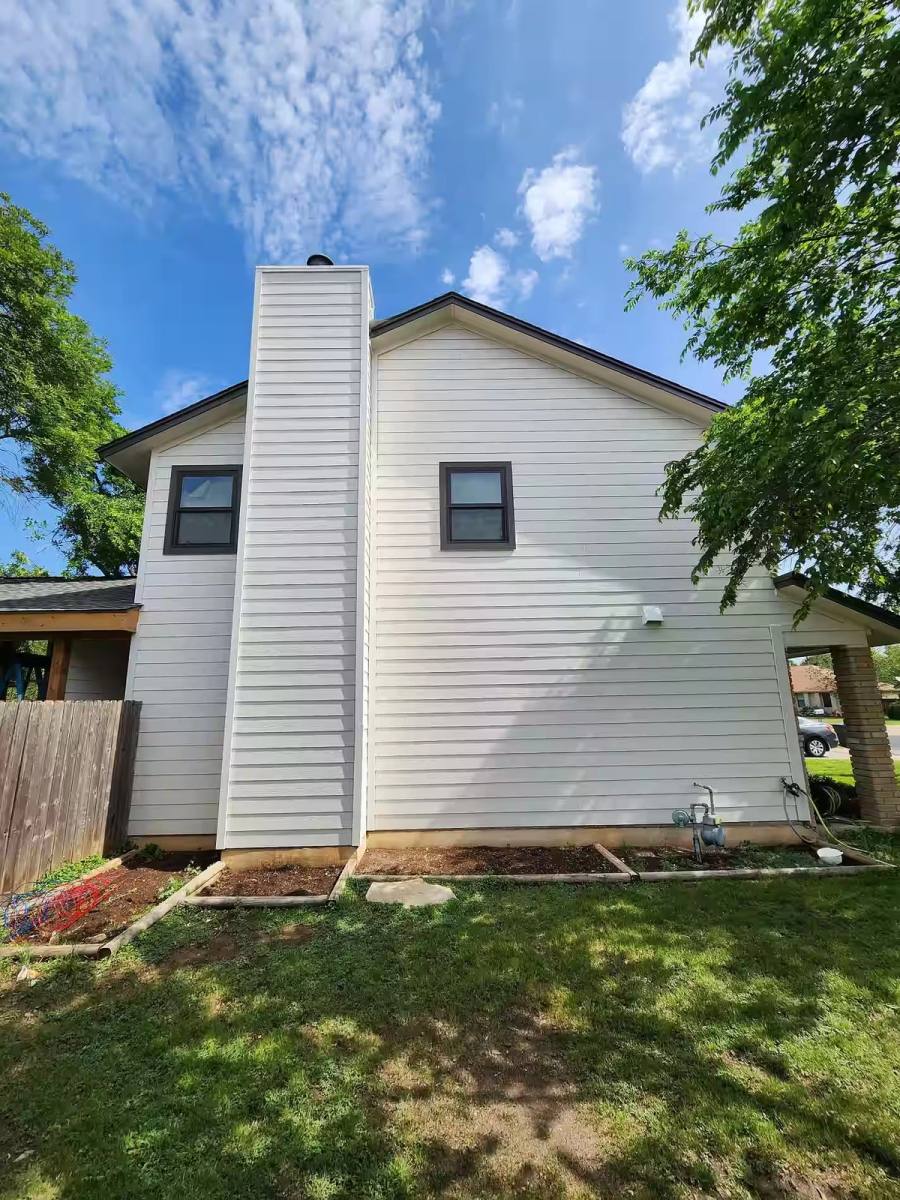The side profile of a residential home renovated with crisp white lap siding and contrasting dark window frames. Features include a siding-clad chimney chase and newly prepared landscaping beds.