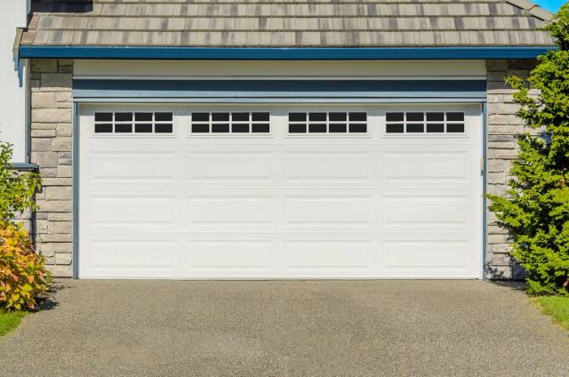 A white garage door with windows, set against a stone wall and green plants, in bright sunlight