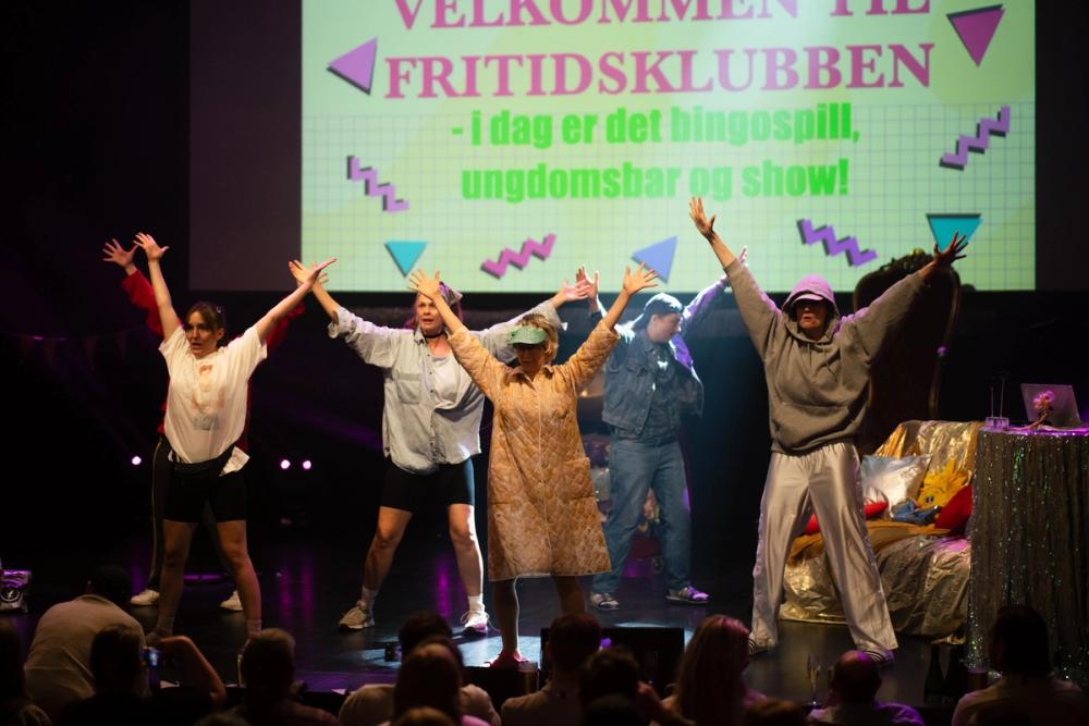 En gruppe mennesker i kostyme danser på en scene med fargerik belysning.