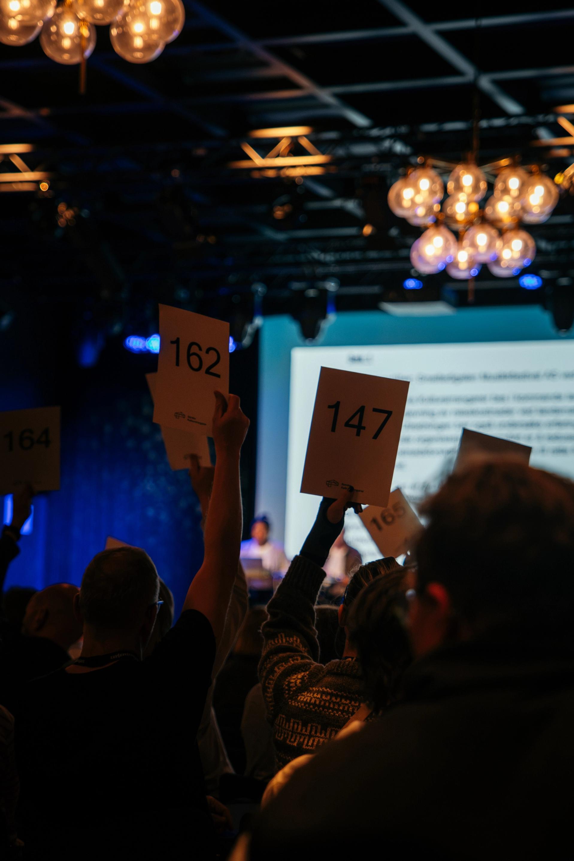 Folk holder opp skilt med numre under en konferanse i et mørklagt lokale med scenelys og runde lamper i taket