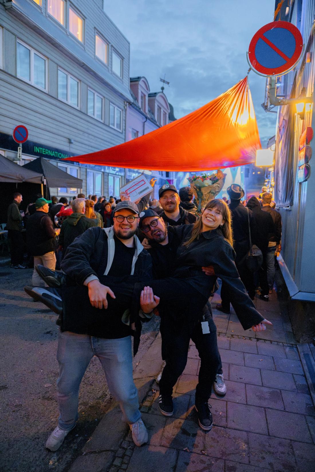 Fire mennesker smiler mot kamera foran et utendørs konsertområde. 