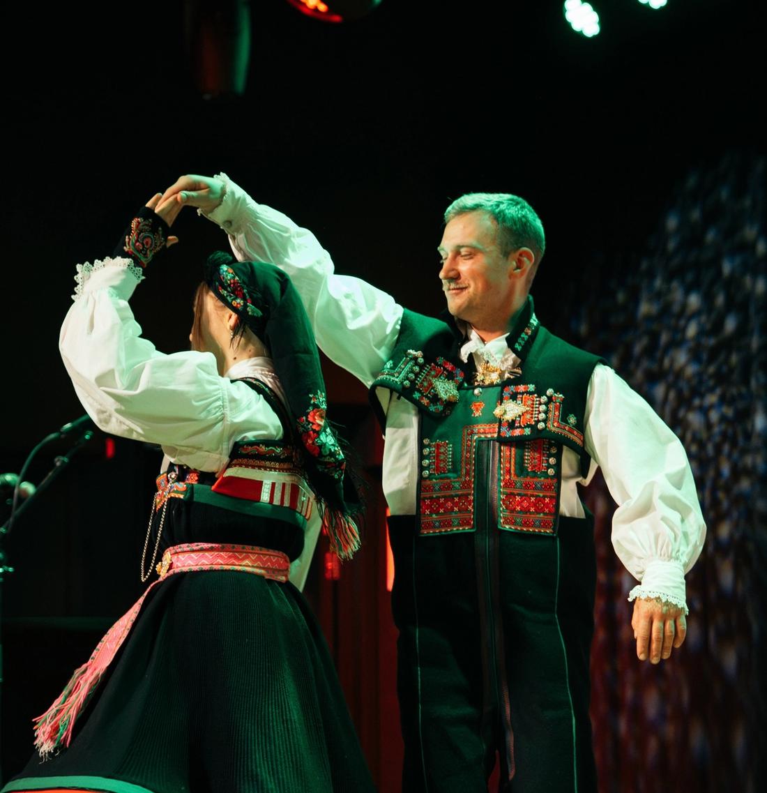 En mann og en kvinne i tradisjonelle folkedrakter danser på en scene.