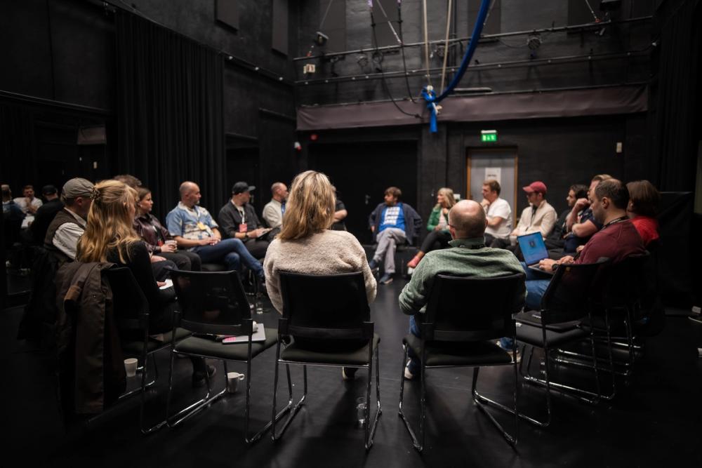 En gruppe mennesker sitter på stoler i en ring og snakker sammen i en black box