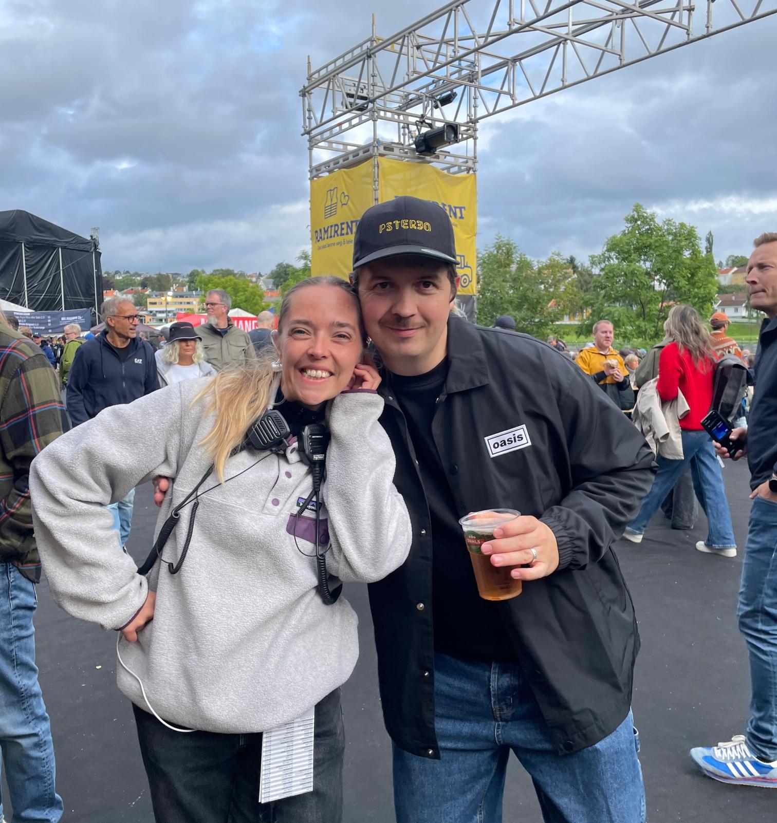 En kvinne og en mann smiler til kamera på et festivalområde.
