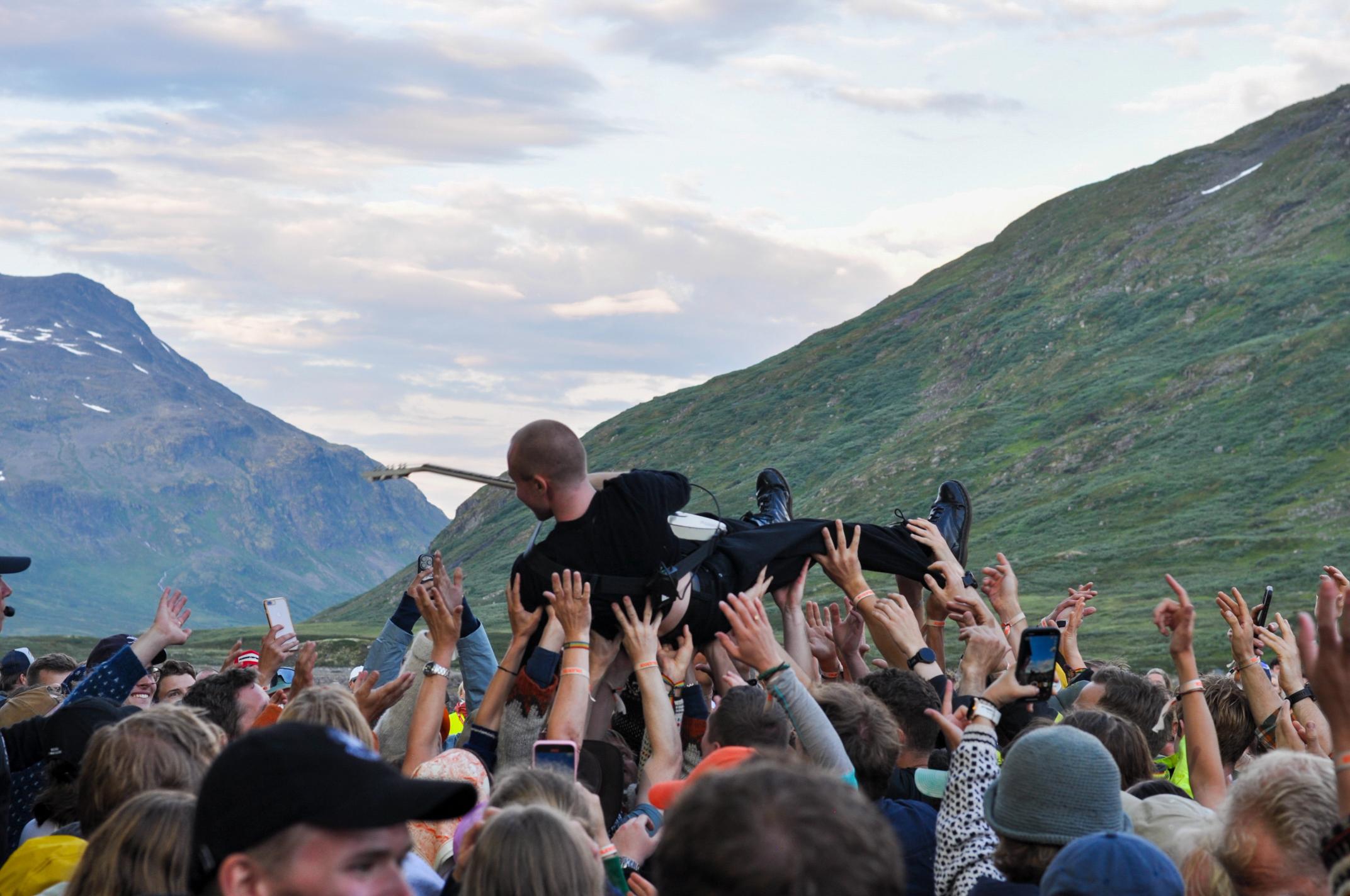 En mann med gitar crowdsurfer på en folkemengde, med mektig fjellandskap i bakgrunnen.