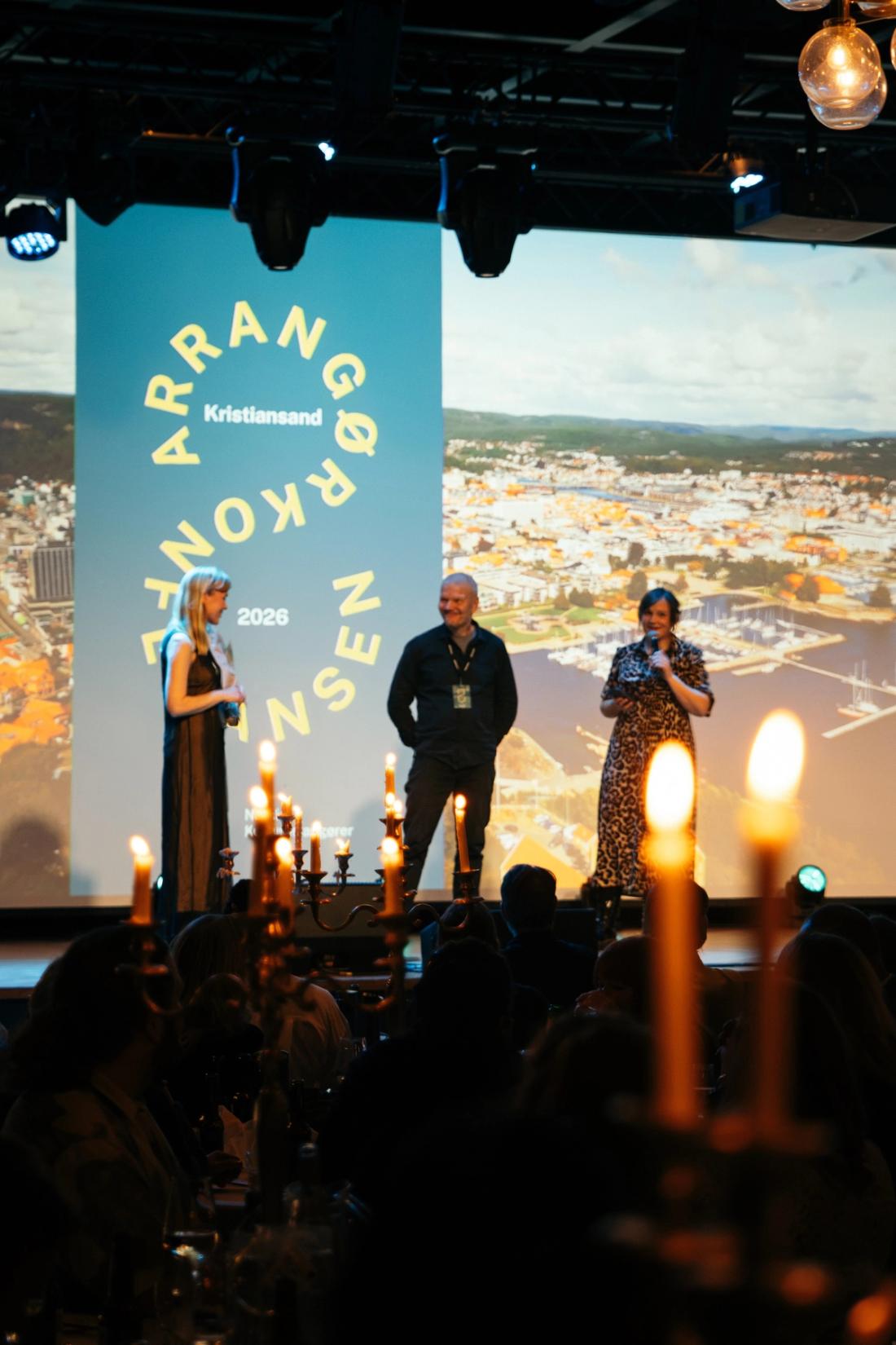 Tre personer står på en scene i et mørkt rom med levende lys på bordene.