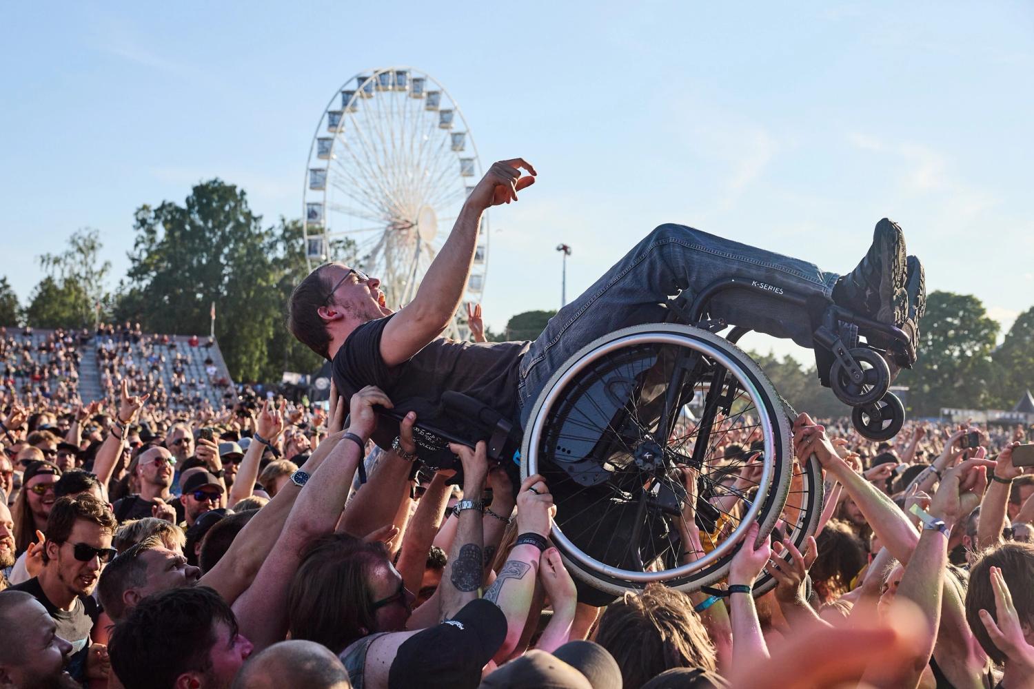 Festivalpublikum løfter en person i rullestol over hodene sine. I bakgrunnen står et pariserhjul.