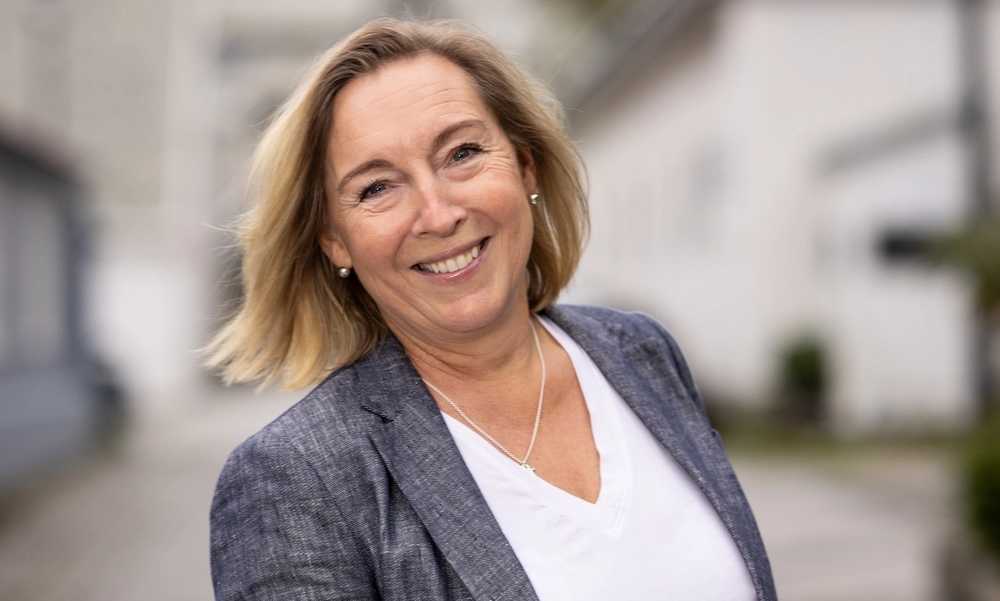 En kvinne med kort lyst hår, hvit tskjorte og grå blazer som smiler med tennene mot kamera.