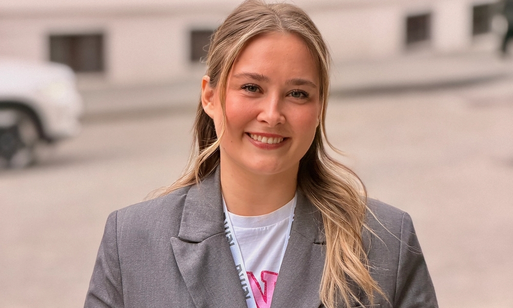 Kvinne med grå dressjakke og t-skjorte med rosa bokstav står med blurry bygate i bakgrunnen.