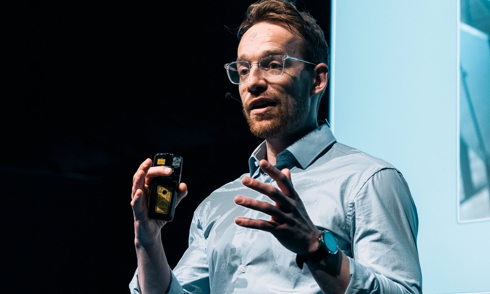 ung mann med briller i ført lyseblå skjorte og sorte bukser står på en scene og foreleser