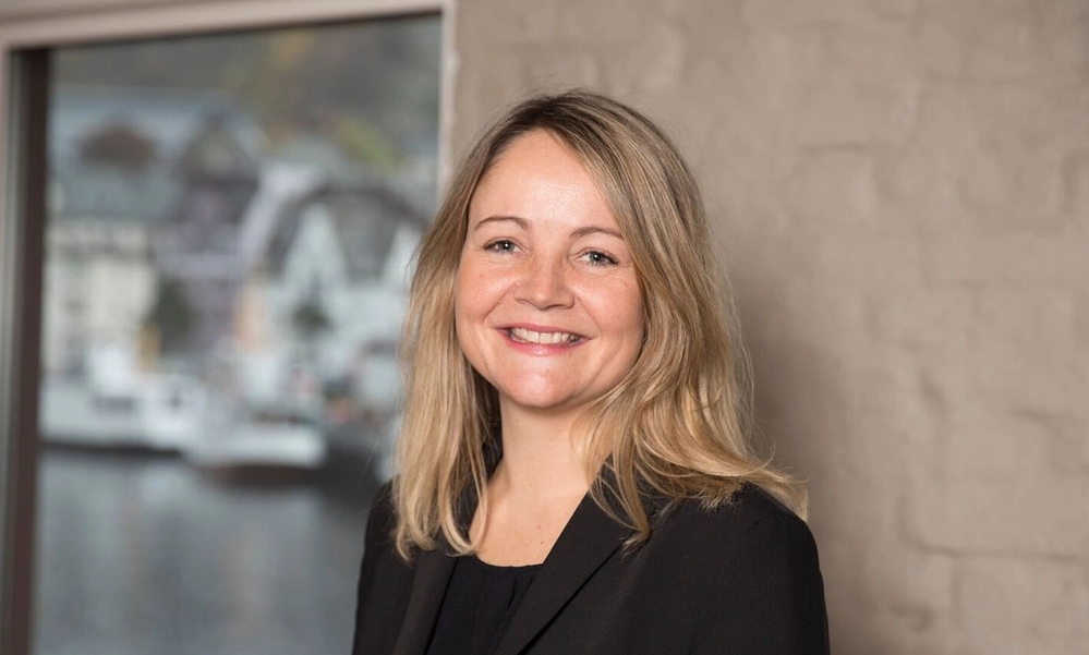 Kvinen smiler med vindu og murvegg i bakgrunnen.