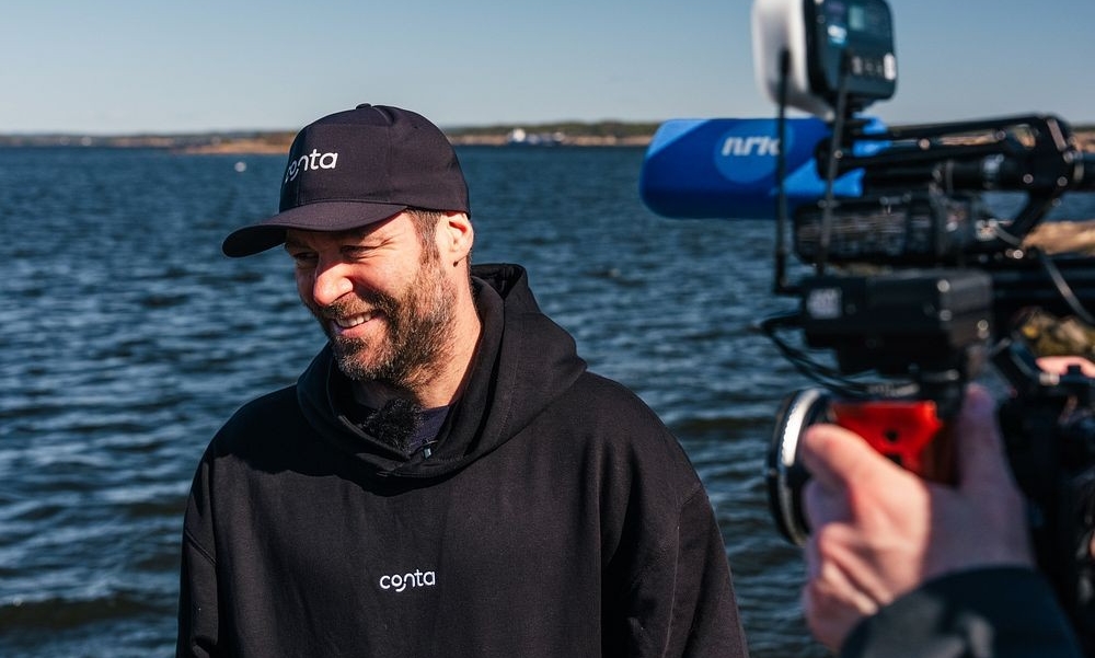 Mann med caps og sort hettegenser står med sjøen i bakgrunnen og et kamera på siden i forgrunnen med NRK-logo på mikrofonen.