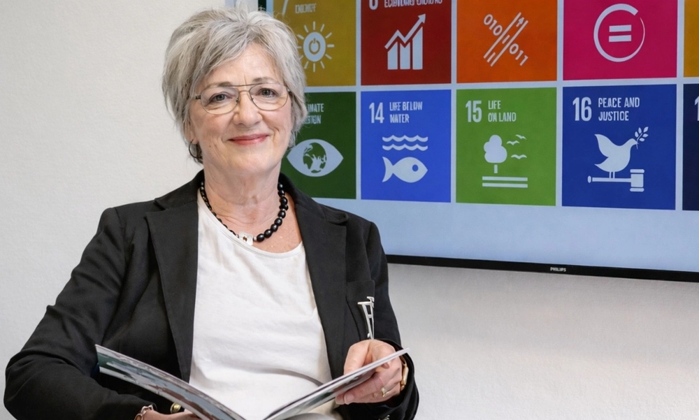 Kvinne med grått hår, dressjakke og hvit topp hår holder bok i fanget