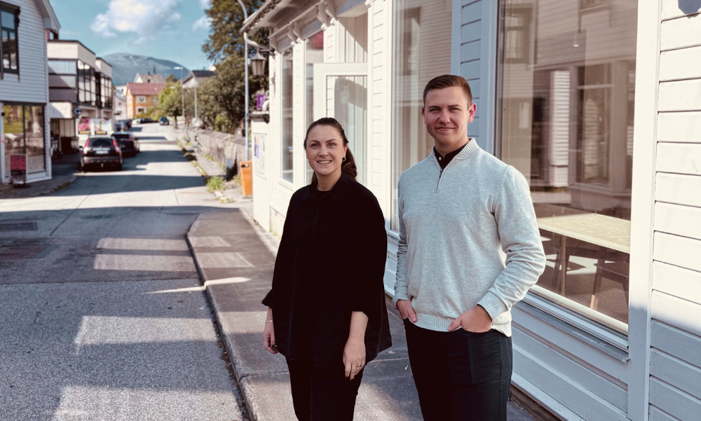 En kvinne og en mann står utenfor på et fortau med hvitt trehus med store vinduer og en gate/bilvei i bakgrunnen.