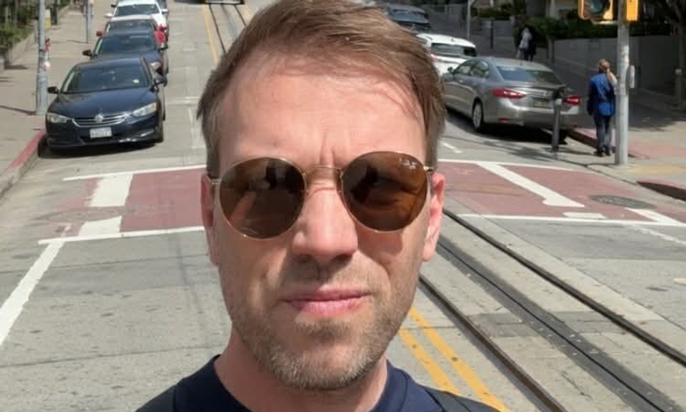 Mann med solbriller og mørkeblå t-skjorte tar selfie i storby.