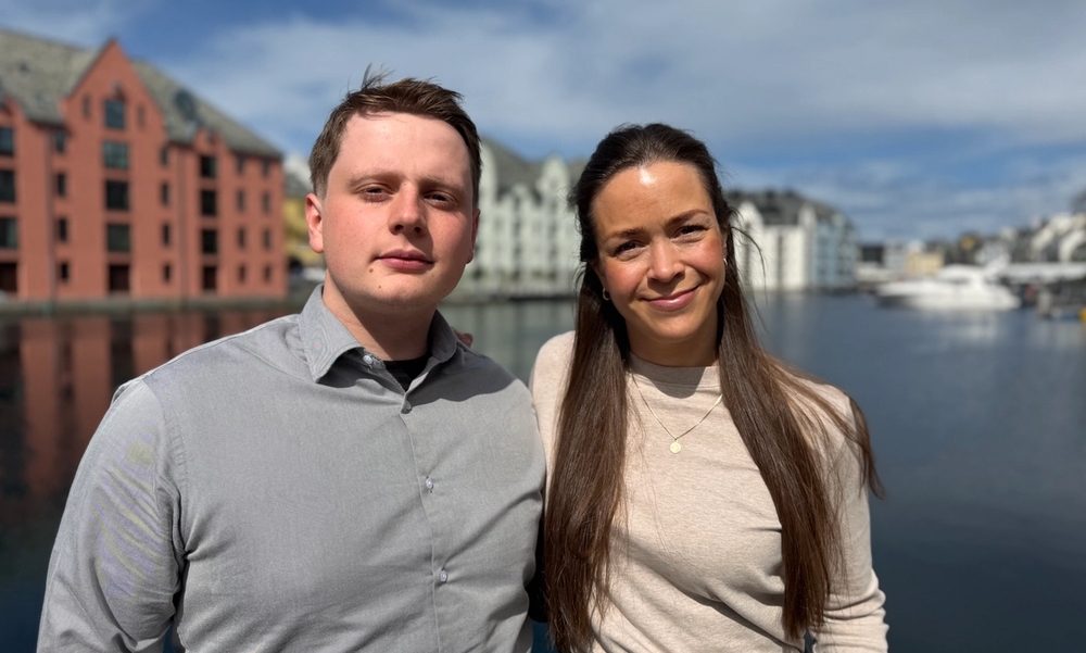 Mann i grå skjorte og kvinne i beige genser smiler, står med sjø og fargerike murbygninger i bakgrunnen.