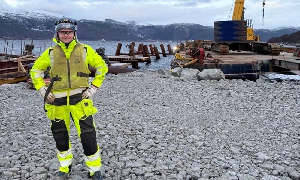 Mann i gult arbeidstøy står på steinmasse med hav og fjell samt heisekran i bakgrunnen