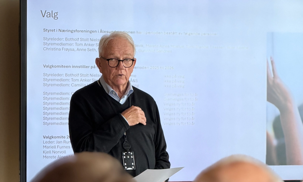 Mann med briller og hvitt hår står foran en stor tv-skjerm med et papir i hånden og prater foran et publikum