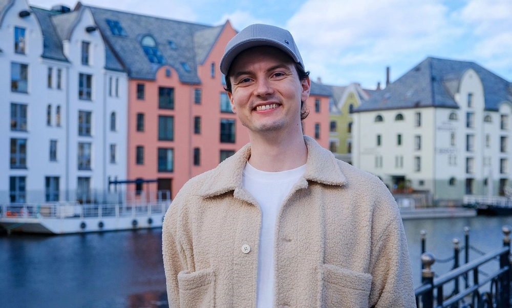 Mann med offwhite teddy-skjorte og blå caps står foran sjø med fargerike hus i bakgrunnen