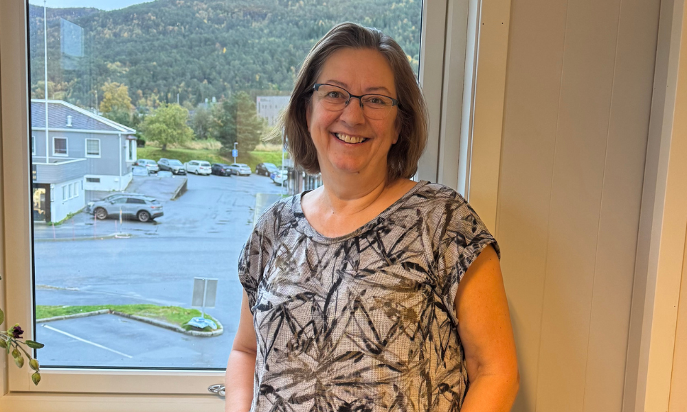 Kvinne med briller står foran et vindu og smiler.