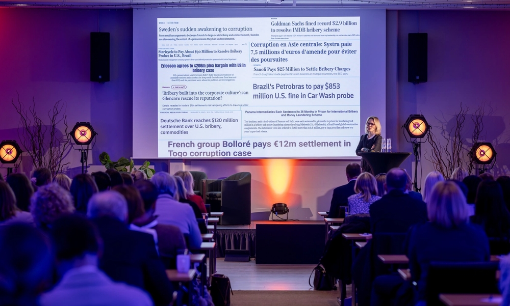 Kvinne står på en scene med presentasjon i bakgrunnen og publikum foran i salen