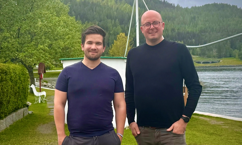 To menn står ved siden av hverandre med hendene i lomma og smiler. Grønt gress og grønne trær i bakgrunnen.