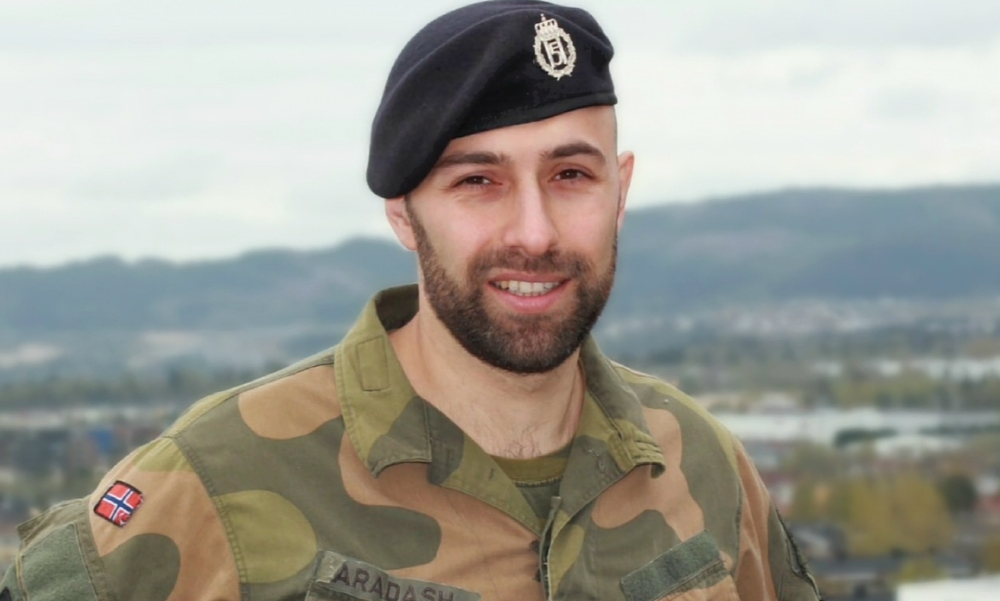 Mann med militæruniform og beret smiler mot kamera med fjell og bygninger i bakgrunnen.