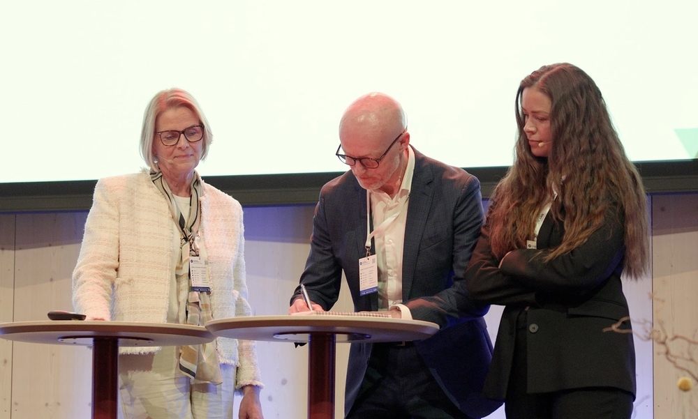 Tre personer står på scene med ståbord. En person signerer et dokument.