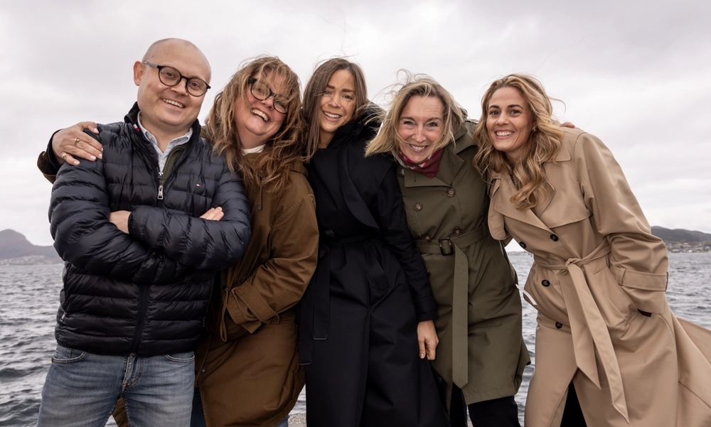 Én mann og fire kvinner står med havet i bakgrunnen, og det blåser.