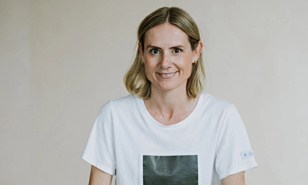 Kvinne i hvit t-skjorte og sort bukse sitter på en pult med en lampe over hodet