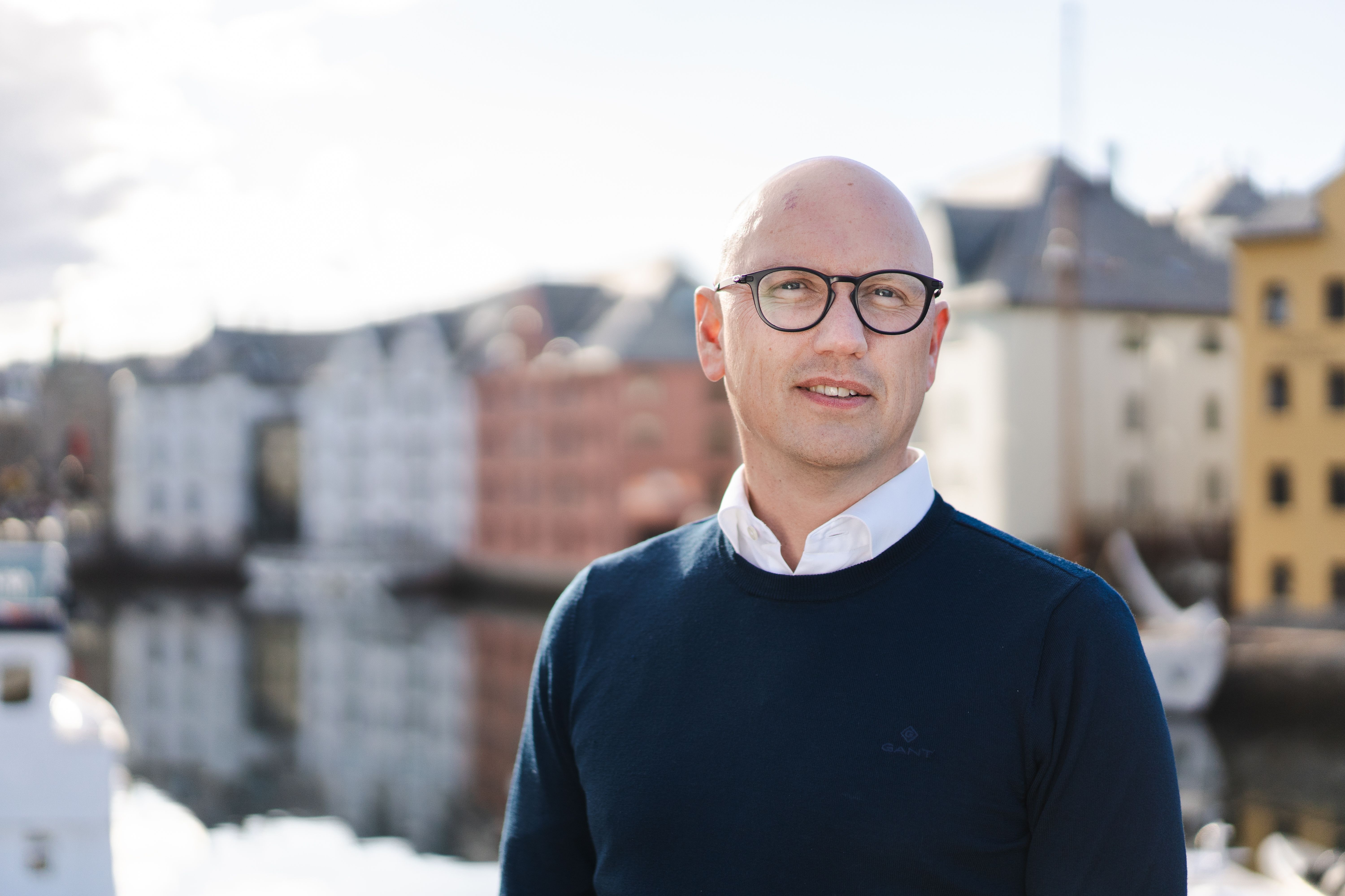 Mann med blå genser, hvit skjorte og briller, uten hår står med blurry fargerike murbygninger og sjø i bakgrunnen.