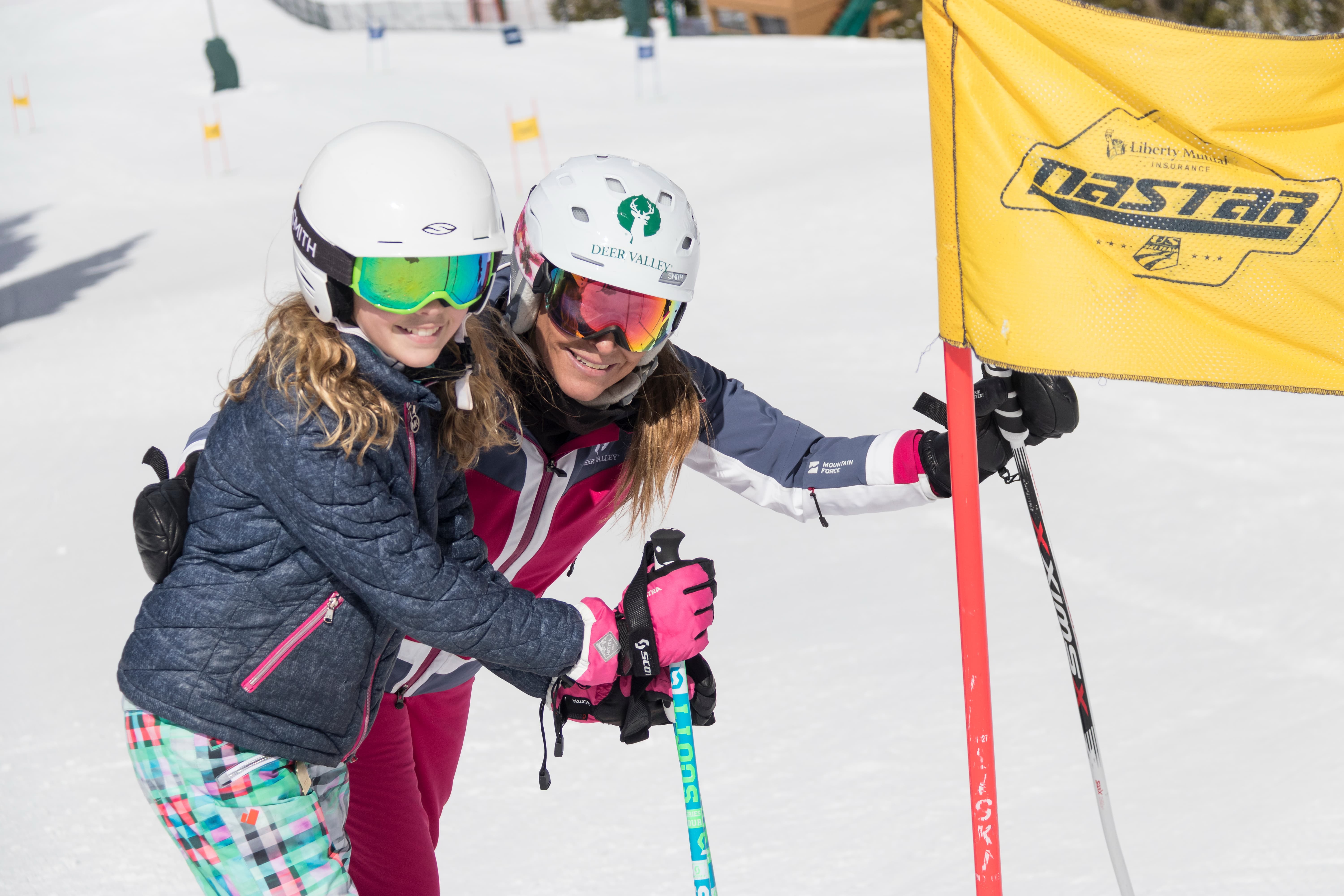 An adult and kid in ski gear standing next to a Nastar ski gate smiling at the camera