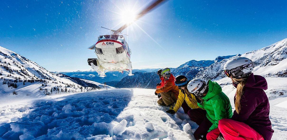 Four people in ski gear watching a helicopter lift off from a snowy mountain top
