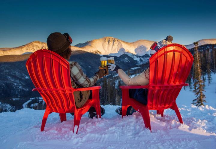 Two people cheers over beers while sitting in adirondack chairs and taking in a mountain view.