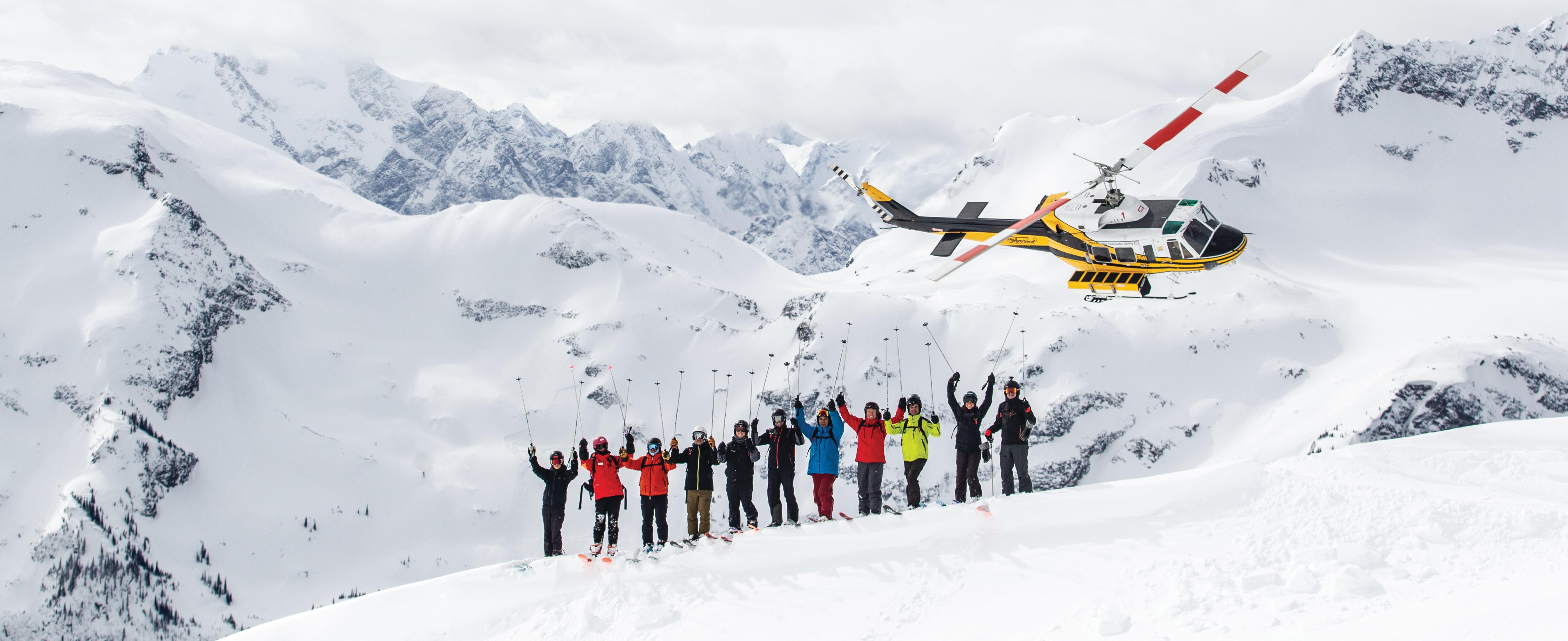 Group of skiers standing in front of a mountain range and a helicopter flying off behind them