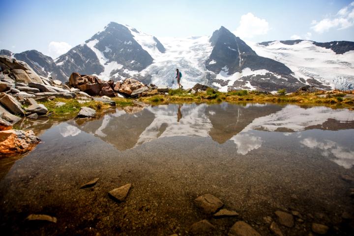 Scenic summer image of a hiker beside a lake with snowy mountains in the background.